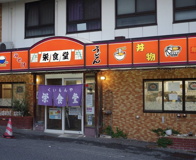 朱色の看板に「栄食堂」、入口に紫の「くいもんや 栄食堂」の暖簾が下がる店構え